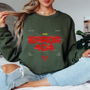 Puede incluir: Una sudadera verde oliva con el texto "ERROR 404" en rojo, con las palabras "OFFLINE", "TIMEOUT", "TRY AGAIN" y "NOT FOUND" alrededor del texto principal. La marca "KINJI" está arriba.