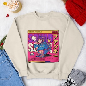 May include: A cream-coloured sweatshirt with an anime-style graphic of a girl with purple hair, smoking, and wearing a backpack. The graphic includes the text "THE HARAUJKU GIRL!" and "SQUAD". The sweatshirt is shown with blue jeans and winter accessories.