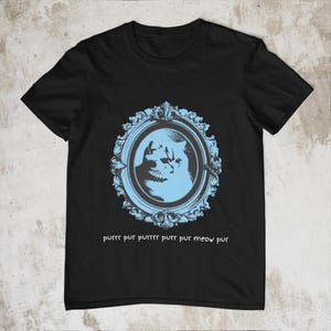 Puede incluir: Camiseta negra con un retrato de gato azul claro en un marco adornado. El diseño incluye el texto "purrr pur purrrr purr pur meow pur". La camiseta está hecha de un material suave. Prenda con temática de gatos.