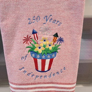 May include: Red and white striped kitchen towel with an embroidered design. The design features a flower pot with yellow flowers, fireworks, and the text "250 Years of Independence" in blue.