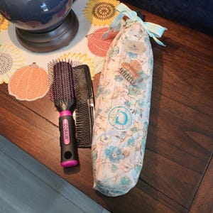 Op de afbeelding: Een zwarte en roze Conair haarborstel met paarse borstelharen, een zwarte kam en een bloemenpatroon tasje vastgebonden met een blauw lint. Een kleine haarclip is aan de tas bevestigd. De tas heeft een logo met de tekst "PAUL & SWEET".
