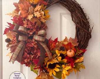 May include: A fall-themed wreath with a grapevine base, adorned with sunflowers, autumn leaves in shades of red, orange, and yellow, and a brown plaid ribbon. A small pumpkin and pinecones add to the seasonal decor.