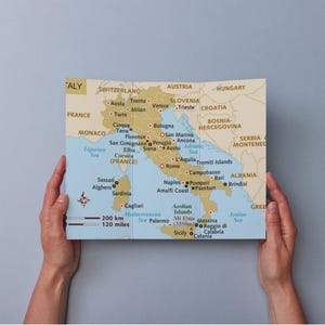 以下が含まれることがあります： 両手で持たれた、イタリアと周辺国の折り畳み地図。地図には都市、水域、200km / 120マイルの縮尺が示されています。地図はタン色で、青と赤の文字が書かれています。