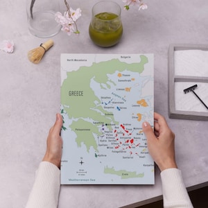 May include: A detailed map of Greece, showing the islands and regions, held by hands. A matcha whisk, a glass of green drink, and flowers are in the background. The map includes a scale of 50 km and 31 miles.