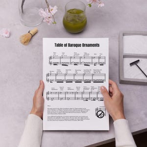 Könnte beinhalten: Ein Blatt Papier mit dem Titel "Tabelle der Barockornamente" mit Noten. Das Papier wird von Händen gehalten, mit einem Glas grüner Flüssigkeit, einem Schneebesen und einem dekorativen Tablett im Hintergrund.