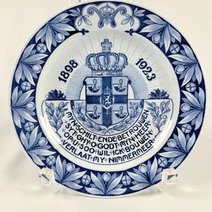 May include: A white and blue decorative plate with a floral border and a central crest design. The plate features text and the years 1898 and 1923. The plate is displayed on a clear stand.