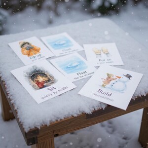 Puede incluir: Un conjunto de tarjetas ilustradas con actividades de invierno, como "Cierra tu chaqueta" y "Construye un mu&ntilde;eco de nieve". Las tarjetas est&aacute;n dispuestas sobre una superficie de madera cubierta de nieve, con un fondo nevado.