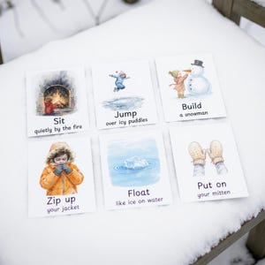 Puede incluir: Seis tarjetas ilustradas con actividades de invierno: sentarse junto al fuego, saltar sobre charcos helados, construir un mu&ntilde;eco de nieve, subir la cremallera de la chaqueta, flotar sobre el hielo y ponerse los guantes. Las tarjetas est&aacute;n sobre una superficie de madera nevada.