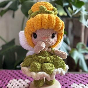 May include: A handmade crochet fairy doll with yellow hair, a green dress, and white wings. The doll has a round face with black eyes and a small nose. It is standing on a wooden base. The background is blurred with green plants.