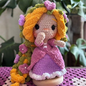 May include: A handmade crochet doll with a light skin tone, wearing a pink and purple dress. The doll has long, orange yarn hair adorned with green leaves and purple flowers. The doll is sitting on a wooden base.