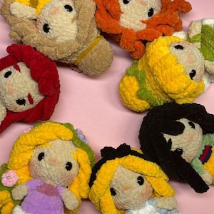 Puede incluir: Una colección de muñecas de ganchillo hechas a mano con varios colores de cabello, incluyendo amarillo, rojo y negro. Las muñecas tienen ojos negros de botón y visten vestidos de diferentes colores. Las muñecas están dispuestas sobre una superficie rosa.