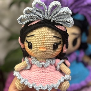 以下が含まれることがあります： 黒い髪、ピンクのドレス、灰色のリボンが付いた手作りのかぎ針編み人形。人形は、黒い目と小さなオレンジ色の口を持つ日焼けした顔をしています。背景には、別の同様の人形が見えます。