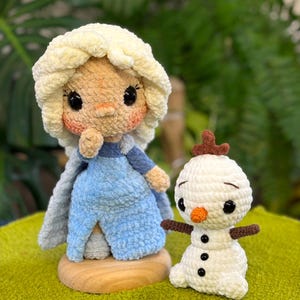 Puede incluir: Una muñeca de ganchillo con cabello de lana amarillo pálido, vestido azul claro y capa a juego, sobre una base de madera redonda. A su lado hay un muñeco de nieve de ganchillo más pequeño en color blanco con nariz naranja.