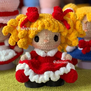 Puede incluir: Una muñeca de ganchillo con cabello rizado amarillo brillante, dos lazos rojos y ojos negros. La muñeca viste un vestido rojo con cuello y dobladillo de volantes blancos, y zapatos negros. El texto @alrevees es visible en el cabello y vestido.
