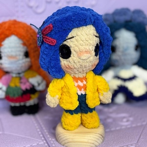 Puede incluir: Muñeca de ganchillo hecha a mano con pelo azul, grandes ojos negros de botón y una chaqueta amarilla. La muñeca está sobre una base de madera, con otras dos muñecas en el fondo. La muñeca tiene un pequeño accesorio para el pelo de libélula.