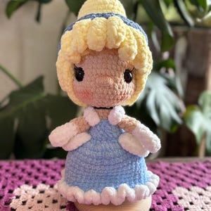 May include: A handmade crochet doll featuring a light blue dress, blonde hair, and a yellow and blue headpiece. The doll has a round face with black eyes and rosy cheeks. The doll is sitting on a wooden base.