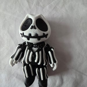 May include: A 3D-printed figurine of a character in a black and white striped suit with a skull-like face. The figure has a white head with black eye sockets and a black mouth. The suit has white stripes on a black background.
