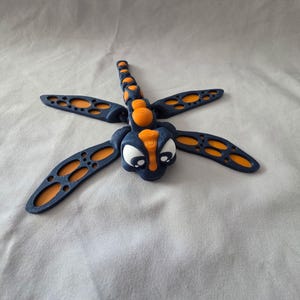 Könnte beinhalten: Eine skurrile Libellenfigur in Marineblau und Orange. Die Libelle hat große, durchbrochene Flügel mit orangefarbenen Akzenten und einen segmentierten Körper. Der Kopf hat große weiße Augen und orangefarbene Details, was ihm ein cartoonartiges Aussehen verleiht.