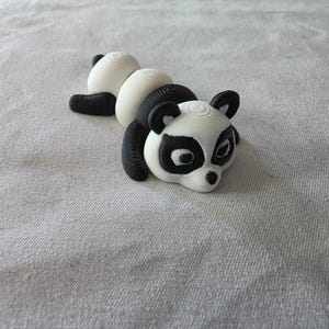 Puede incluir: Figura de panda impresa en 3D en posición tumbada. El panda tiene un esquema de color blanco y negro, con orejas, patas y marcas alrededor de los ojos negras. El cuerpo es blanco con detalles negros, sobre una superficie gris claro.