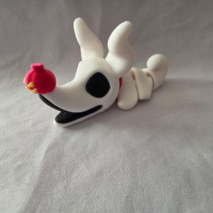 Könnte beinhalten: Eine weiße Zero-Figur aus The Nightmare Before Christmas. Die Figur hat ein schwarzes Auge, eine rote Nase und einen schwarzen Mund. Der Körper ist segmentiert und weiß, mit einem roten Kragen.