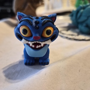 Peut inclure: Une petite figurine de créature bleue imprimée en 3D avec de grands yeux jaunes, des rayures noires sur la tête, un nez rose et une large bouche montrant des crocs blancs. La figurine a une tache bleu clair sur la poitrine et quatre petites pattes.