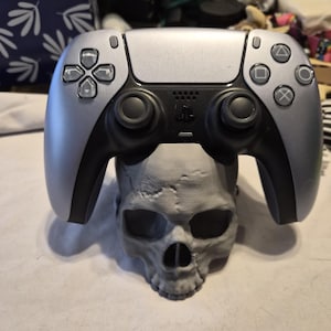 Op de afbeelding: Een zilver-zwarte PlayStation 5-controller rust bovenop een grijze schedel. De controller heeft een symmetrisch ontwerp met zwarte joysticks en knoppen, en een PlayStation-logo. De schedel is een gedetailleerd, driedimensionaal model.