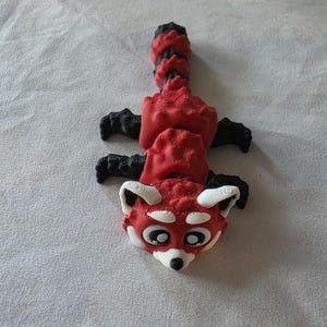 Peut inclure: Figurine de panda roux avec un design détaillé. La figurine présente un corps rouge, des pattes et des anneaux de queue noirs, et un visage blanc et noir. Le panda est allongé sur une surface blanche.