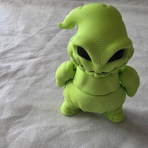 Puede incluir: Una figura verde lima de un personaje con un cuerpo bulboso, brazos y piernas pequeños. La figura tiene una cabeza grande con una parte superior puntiaguda y una sonrisa siniestra. Los ojos son negros y huecos. Toda la figura tiene una apariencia texturizada y en capas.