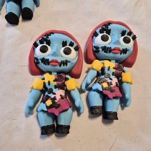 May include: Handmade figurines of a character with blue skin, red hair, and stitched features. The character wears a patchwork dress with yellow, pink, and black accents. Black boots complete the look. The figurines are likely inspired by a popular animated film.