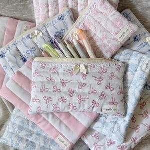 May include: A collection of quilted fabric pencil cases and pouches in various patterns. The cases feature white fabric with pink bow patterns, pink and white striped fabric, and blue floral patterns. Each case has a zipper closure and a small bow.