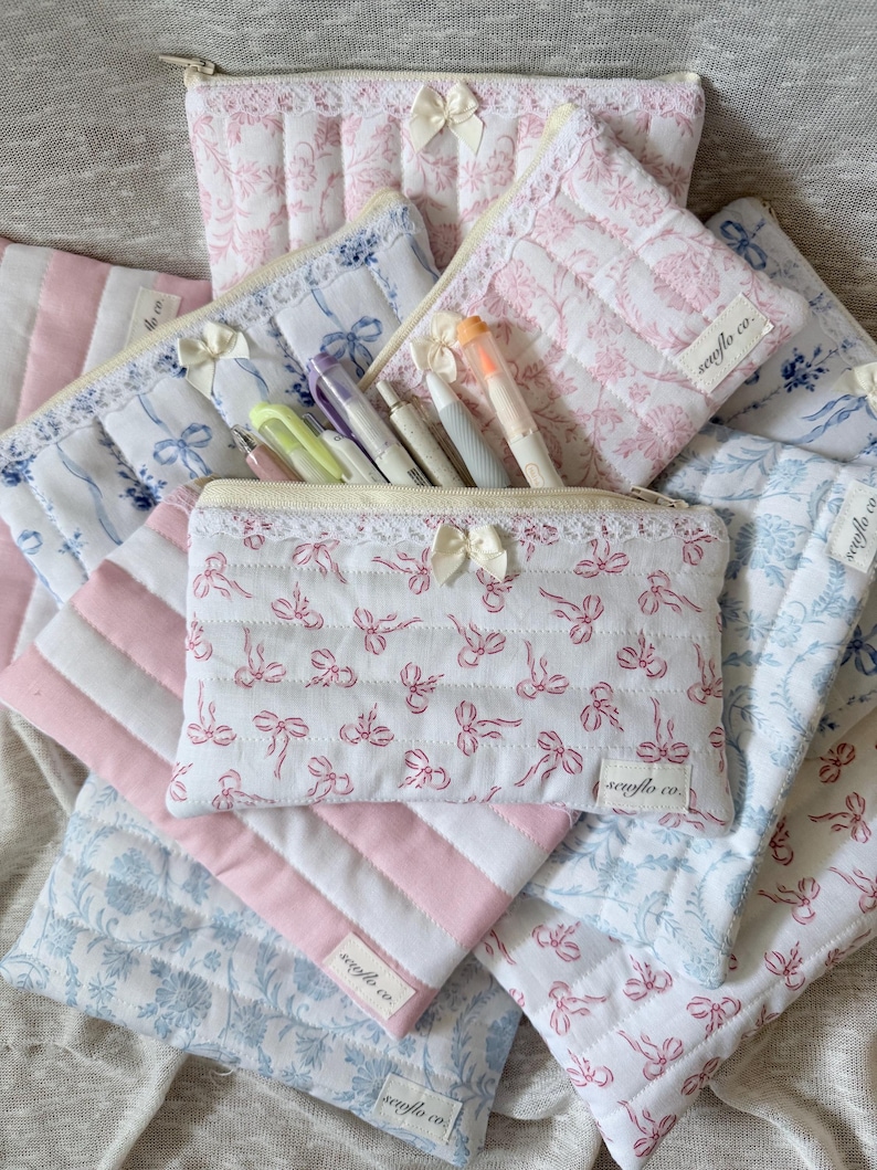 May include: A collection of quilted fabric pencil cases and pouches in various patterns. The cases feature white fabric with pink bow patterns, pink and white striped fabric, and blue floral patterns. Each case has a zipper closure and a small bow.
