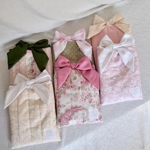 May include: A collection of four rectangular quilted fabric pouches, each featuring a decorative fabric bow at the top. The pouches display various patterns including floral, paisley, and polka dot designs in shades of pink, cream, green, and white.