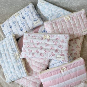 May include: A collection of quilted cosmetic bags in various patterns and colors. The bags feature floral and bow designs in shades of blue and pink, with white lace and ribbon accents. Each bag has a zipper closure.