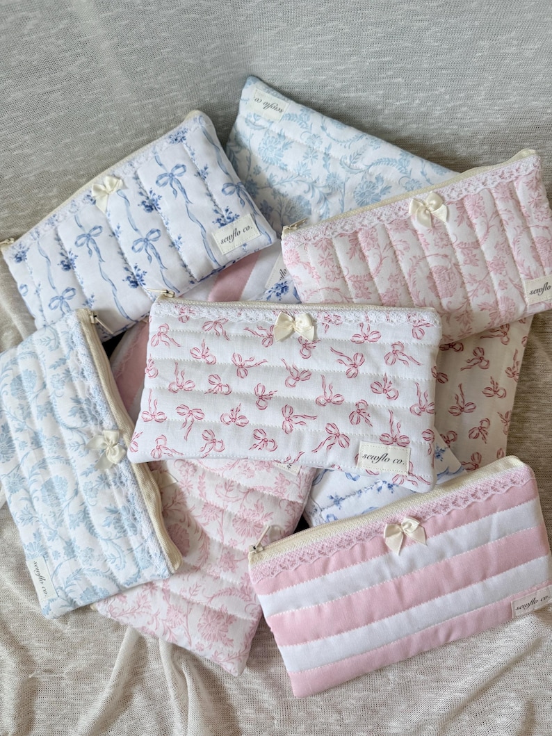 May include: A collection of quilted cosmetic bags in various patterns and colors. The bags feature floral and bow designs in shades of blue and pink, with white lace and ribbon accents. Each bag has a zipper closure.