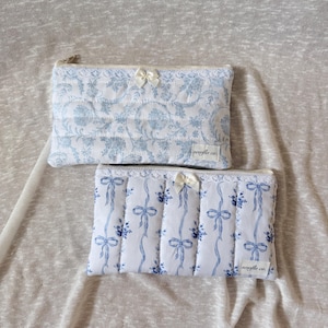 May include: Two quilted cosmetic bags. The top bag features a blue floral pattern on a white background, with a small bow. The bottom bag has a blue ribbon design on a white background, with a small bow. Both bags have a zipper closure.