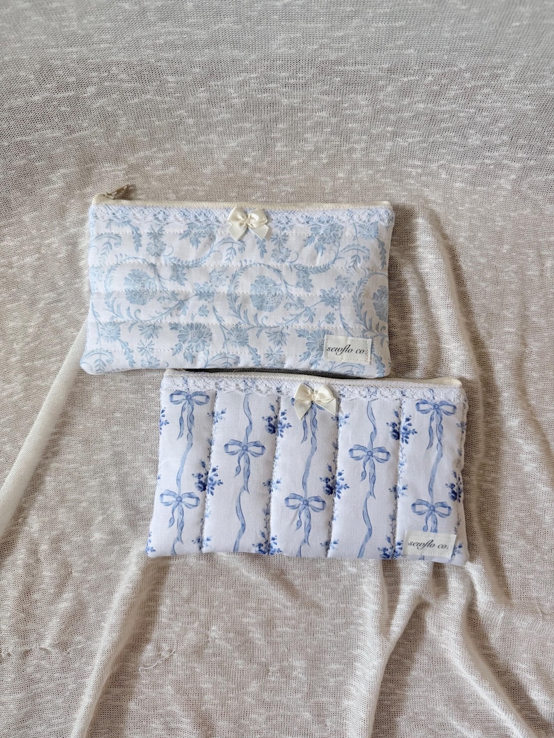 May include: Two quilted cosmetic bags. The top bag features a blue floral pattern on a white background, with a small bow. The bottom bag has a blue ribbon design on a white background, with a small bow. Both bags have a zipper closure.