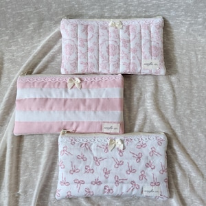 May include: Three quilted cosmetic bags in pink and white. One bag features a floral pattern, another has pink and white stripes, and the third displays a bow pattern. Each bag has a zipper closure, lace trim, and a small bow.
