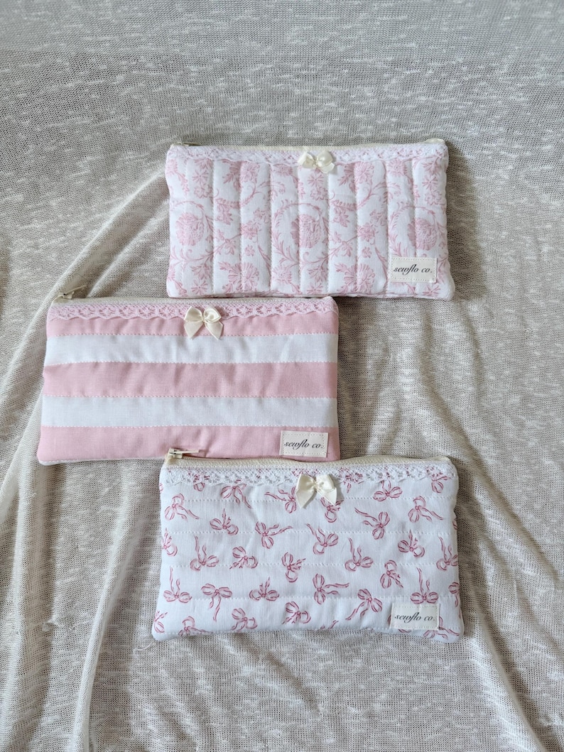 May include: Three quilted cosmetic bags in pink and white. One bag features a floral pattern, another has pink and white stripes, and the third displays a bow pattern. Each bag has a zipper closure, lace trim, and a small bow.