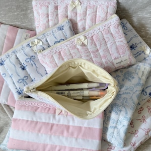 May include: A collection of quilted fabric pouches in various patterns and colors. Some pouches feature pink and white floral designs, while others have blue floral patterns or pink and white stripes. Each pouch has a zipper closure and a small bow.