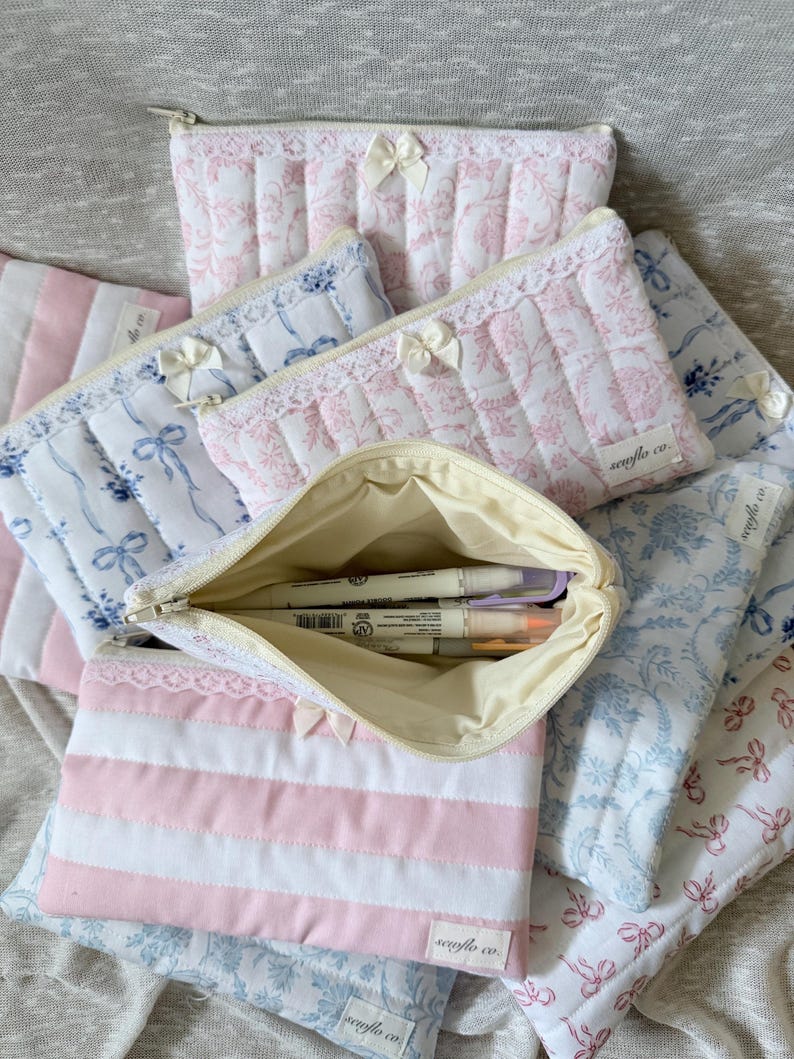May include: A collection of quilted fabric pouches in various patterns and colors. Some pouches feature pink and white floral designs, while others have blue floral patterns or pink and white stripes. Each pouch has a zipper closure and a small bow.