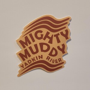May include: A tan sticker with the words "MIGHTY MUDDY YADKIN RIVER" in dark brown. The sticker has a unique shape with wavy lines at the top, suggesting a river theme. The design is simple and bold.