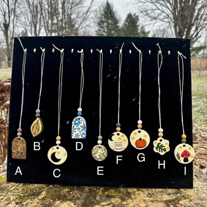 May include: A collection of nine handmade pendant necklaces displayed on a black surface. Each pendant features unique designs, including a leaf, moon, owl, pumpkin, and mushroom. The pendants are strung on white cords with decorative beads.
