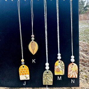May include: A collection of handmade ceramic pendants hanging against a black backdrop. The pendants feature various designs, including a honeycomb, leaf, and floral patterns. Each pendant is suspended by a white cord with decorative beads.