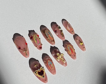 May include: A collection of ten long, almond-shaped artificial nails. Each nail is painted a light pink and adorned with various embellishments, including religious imagery, gold crosses, red gems, and gold heart-shaped charms. Some nails feature small gold studs.