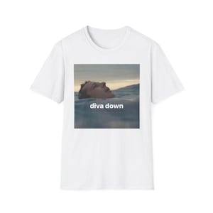May include: White t-shirt featuring a square image of a person's face emerging from water, with the text "diva down" in white. The image has a soft, muted color palette, with blues, tans, and whites.