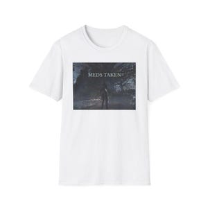 Könnte beinhalten: Weißes T-Shirt mit einem rechteckigen Grafikdesign. Die Grafik zeigt eine dunkle, atmosphärische Szene mit einer Figur in einem Wald und dem Text "MEDS TAKEN" in Weiß. Das T-Shirt hat kurze Ärmel und einen Rundhalsausschnitt.