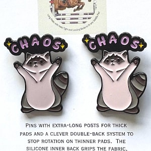 May include: Two enamel pins featuring a raccoon with outstretched arms. Each pin has the word "CHAOS" above it in a bubble letter style. The pins are light pink, gray, and black. A small image of a horse jumping an obstacle is in the background.