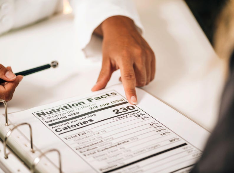 May include: A close-up of a nutrition facts label with the text "Nutrition Facts" at the top. The label shows the number of calories per serving, with a hand pointing to the number 230. A pen is also visible.