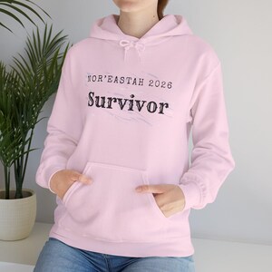 Könnte beinhalten: Ein hellrosa Kapuzenpullover mit einer Vordertasche und einer Kapuze mit Kordelzug. Der Text "NOR'EASTAH 2026 Survivor" ist in Schwarz auf die Brust gedruckt. Die Person trägt eine blaue Jeans.