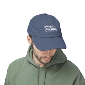 Puede incluir: Gorra de béisbol azul desgastada con la palabra "Survivor" en blanco, junto con el texto "SOMEWHERE 2019". La gorra tiene una visera curva y un aspecto desgastado, lo que sugiere un estilo vintage o robusto.